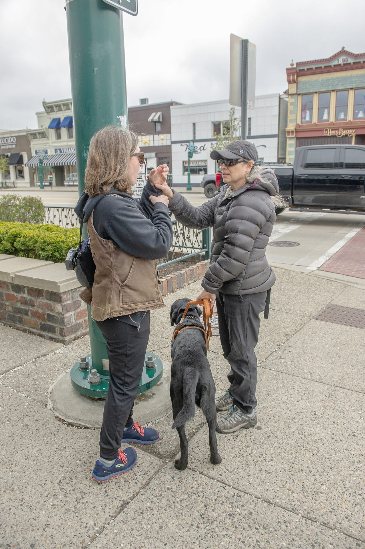 Two people converse on a city sidewalk while a dog stands patiently beside them.