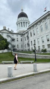 A woman uses her white cane while walking beside a four-story, granite state capital building with a large dome on top.