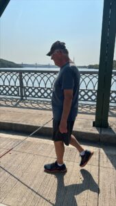 A man walking along a paved pier on a sunny day. HE is using a white cane as he walks.