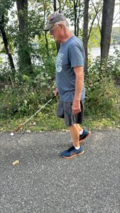 A man walking along a wooded train using a white cane. He is wearing shorts, a t-shirt, and hat.