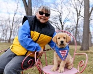 A person wearing sunglasses and a yellow and black vest sits outside in a wooded area. On a chair next to her is a small yellow Labrador puppy wearing a blue Future Leader Dog bandana. 
