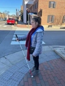 A woman standing on a street corner in winter gear holding a white cane. 