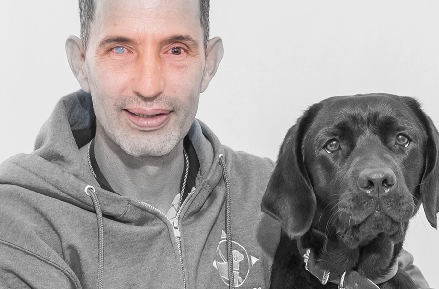 Close-up black and white photo of man smiling slightly next to a black lab. The area just around his eyes is in color, showing that one is light blue and the other one brown