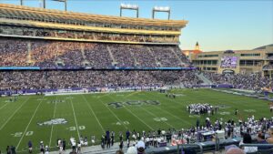 A football stadium full of people. the letters "TCU" is written on the turn in the middle of the field surrounded by players in blue uniforms.