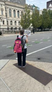 A person wearing a gray shirt and carrying a red and black backpack is holding a sign that reads help me CROSS STREET on a sidewalk in an urban area with historic buildings and trees in the background.