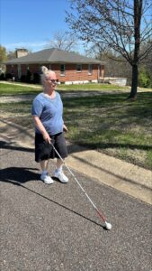 Person using a white cane walking on a sidewalk in front of a brick house, with trees and a clear blue sky in the background.
