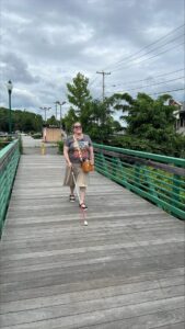 A woman uses her white cane as she walks across a wooden bridge. She is wearing a t-shirt and long skirt and is smiling widely.