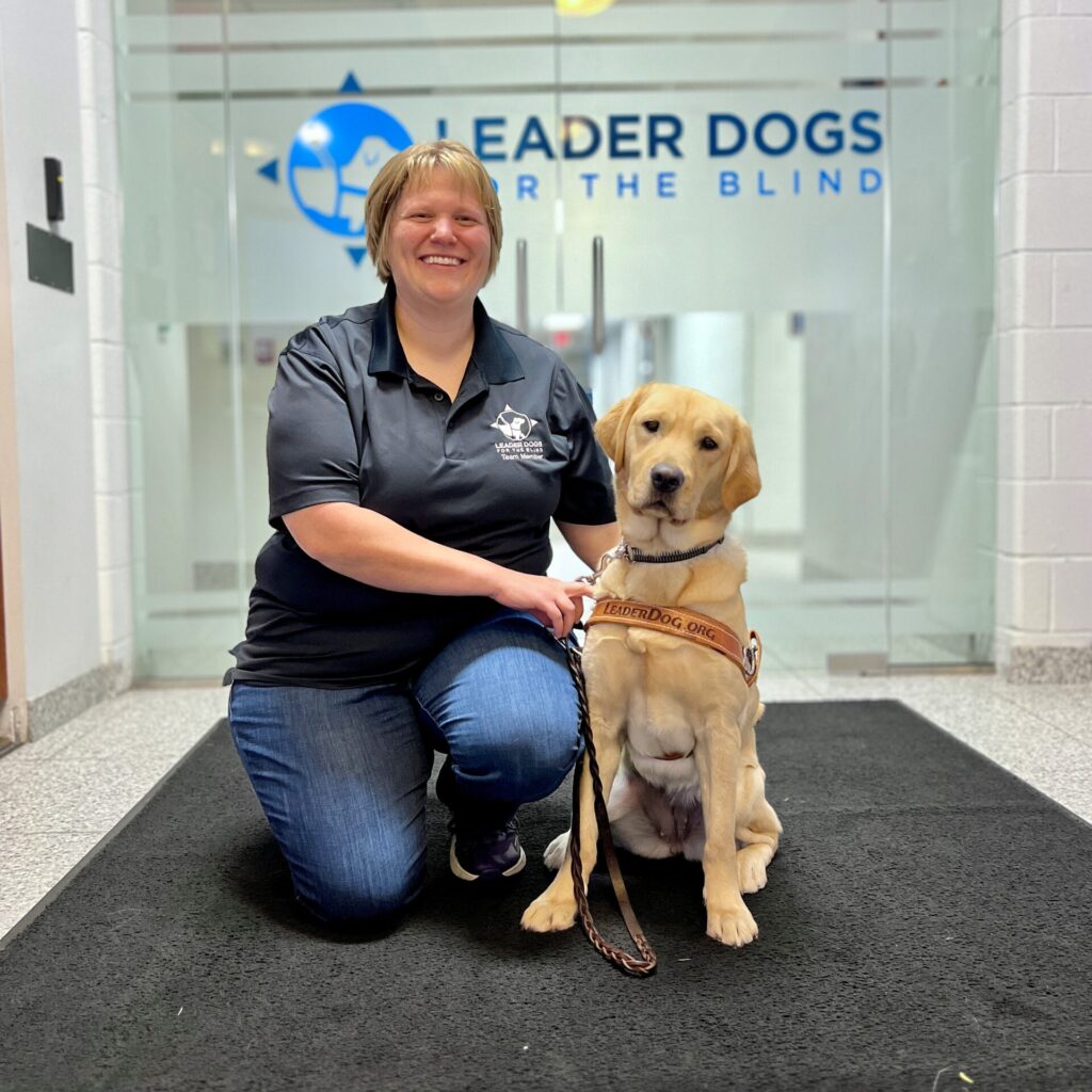 A smiling woman kneels beside a yellow Labrador retriever wearing a harness, in front of a doorway featuring the logo for Leader Dogs for the Blind.