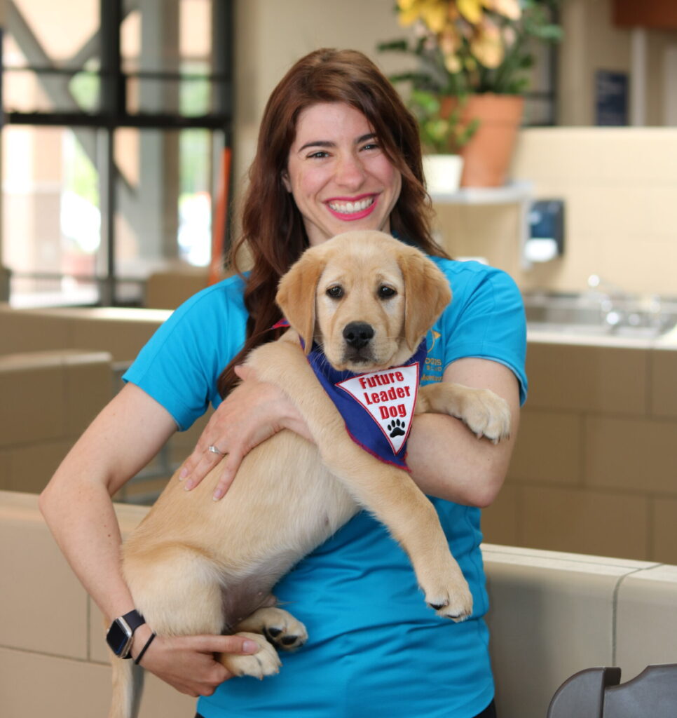 A smiling woman in a blue shirt holds a playful yellow puppy wearing a bandana that reads "Future Leader Dog."