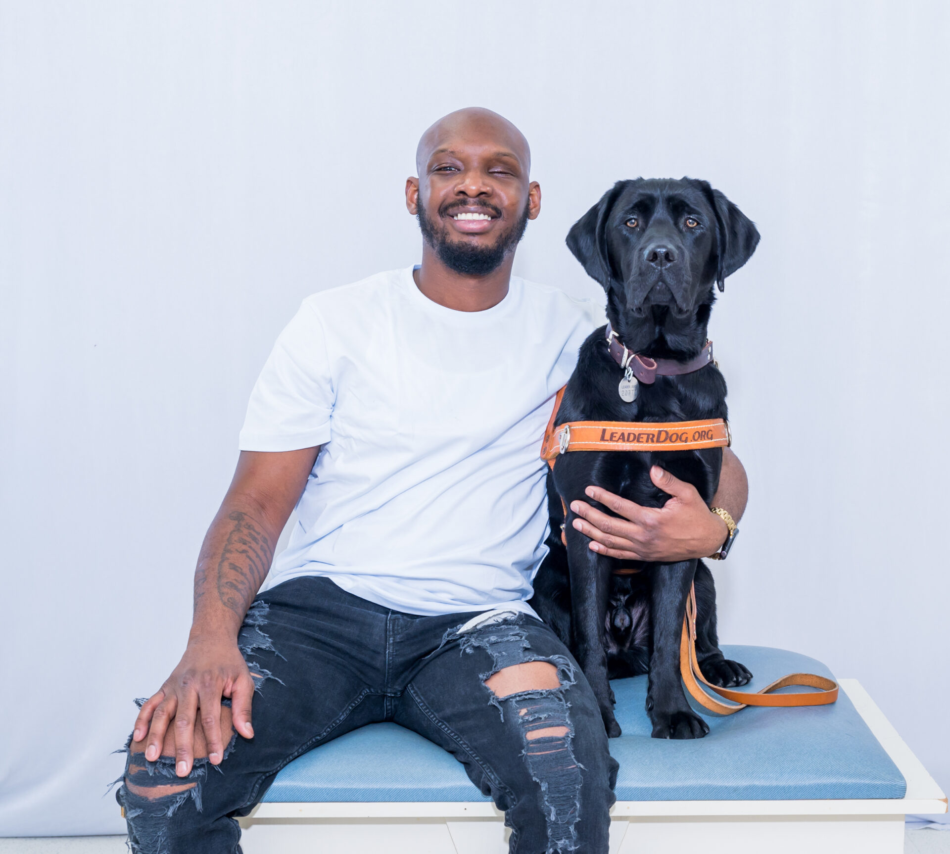 A smiling individual sits on a bench beside a service dog wearing a harness.