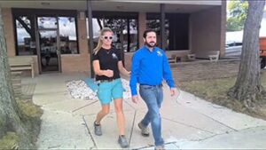 Two individuals are walking together outside a building, with one wearing sunglasses and casual attire.