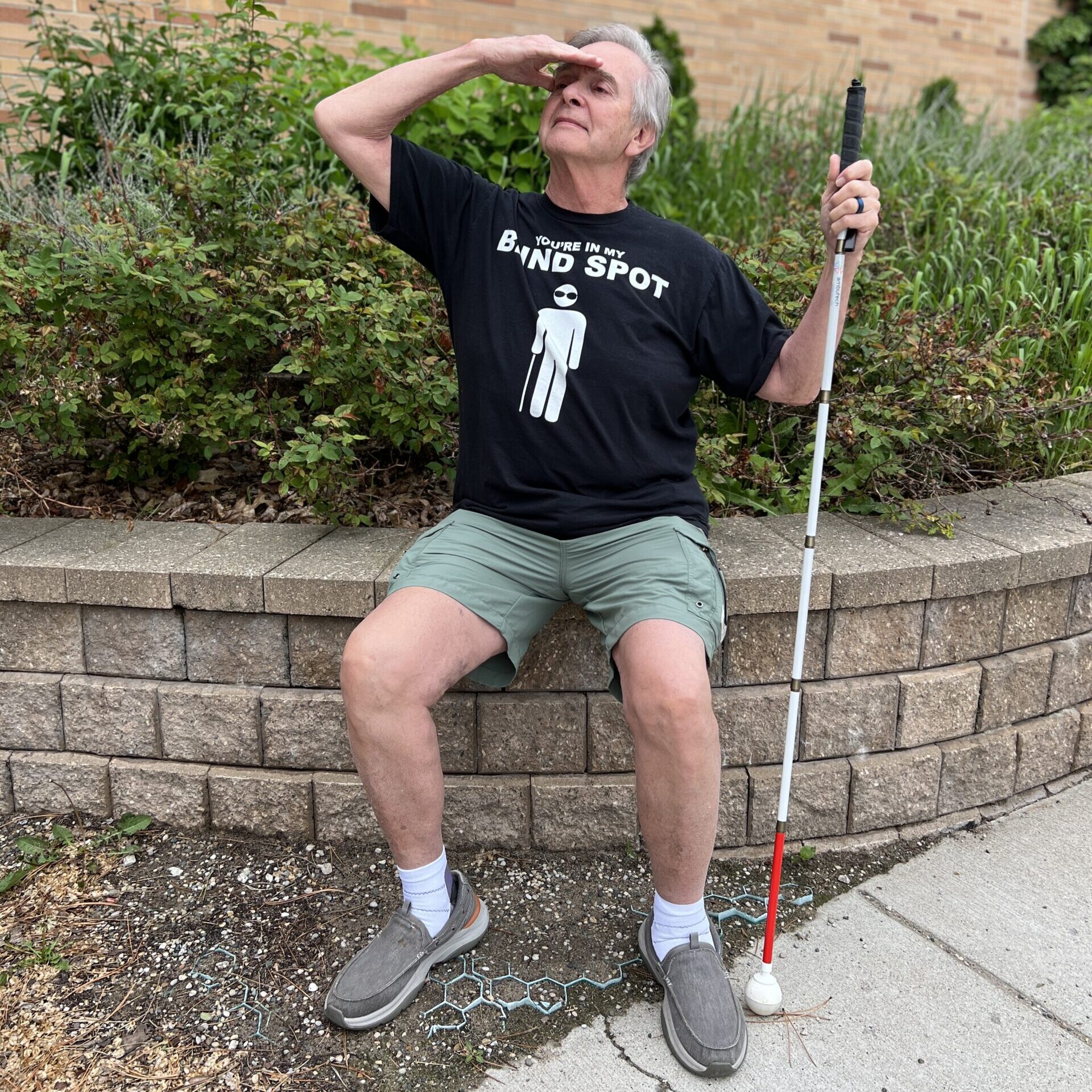 A person wearing a humorous t-shirt sits on a stone wall, shielding their eyes from the sun while holding a white cane.
