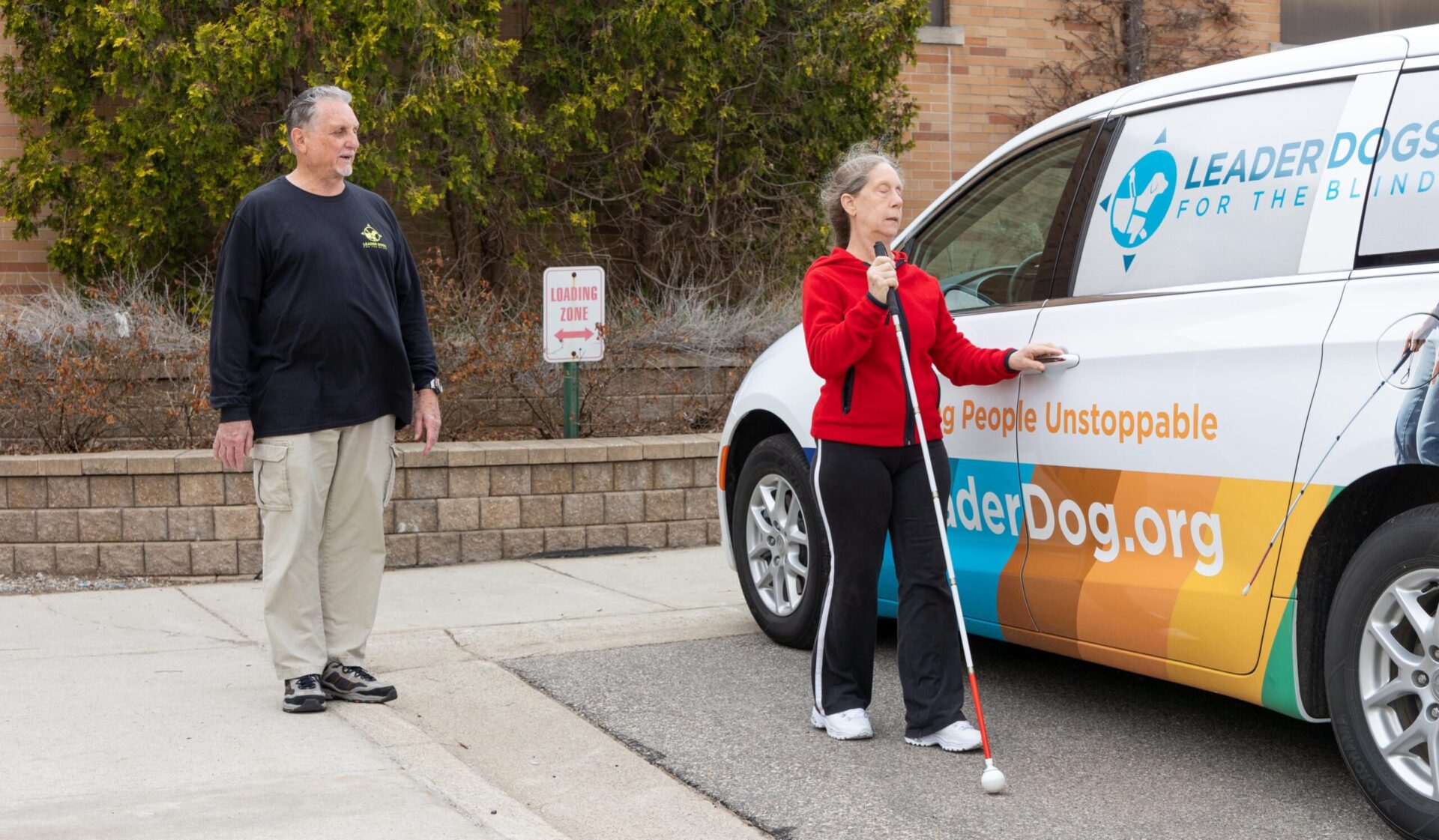 A woman in a red jacket uses a white cane while standing by a vehicle branded for a program supporting individuals with visual impairments.