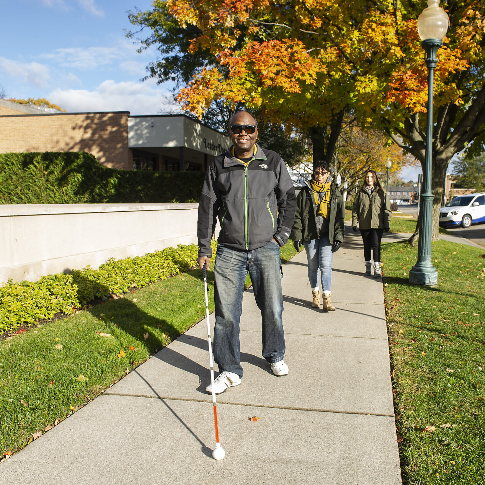 A group of individuals walks along a sidewalk lined with trees displaying autumn foliage.