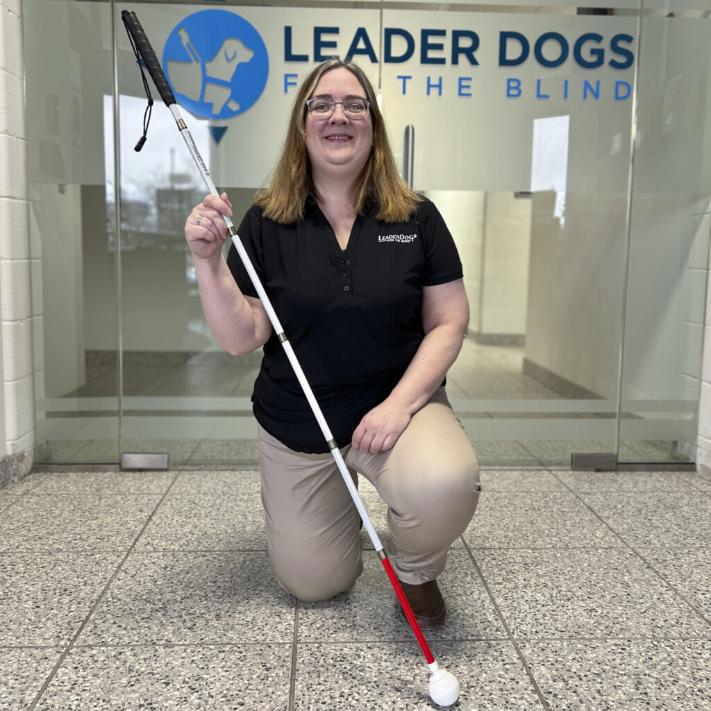 A person is kneeling and smiling while holding a white cane in front of a sign for an organization supporting blind individuals.