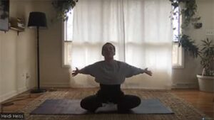 A person is sitting in a yoga pose with arms outstretched, surrounded by natural light and a cozy indoor setting.