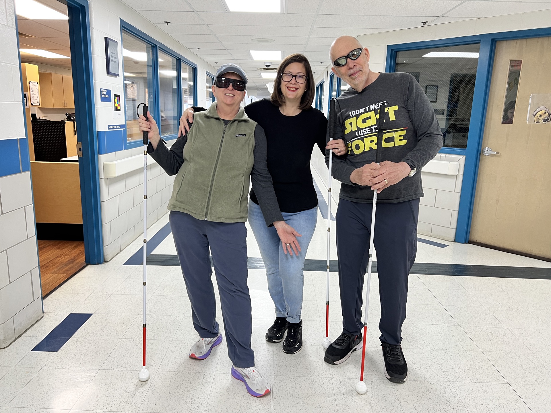Three individuals are standing together in a hallway, each holding a white cane and wearing sunglasses, with one person playfully posing in the center.
