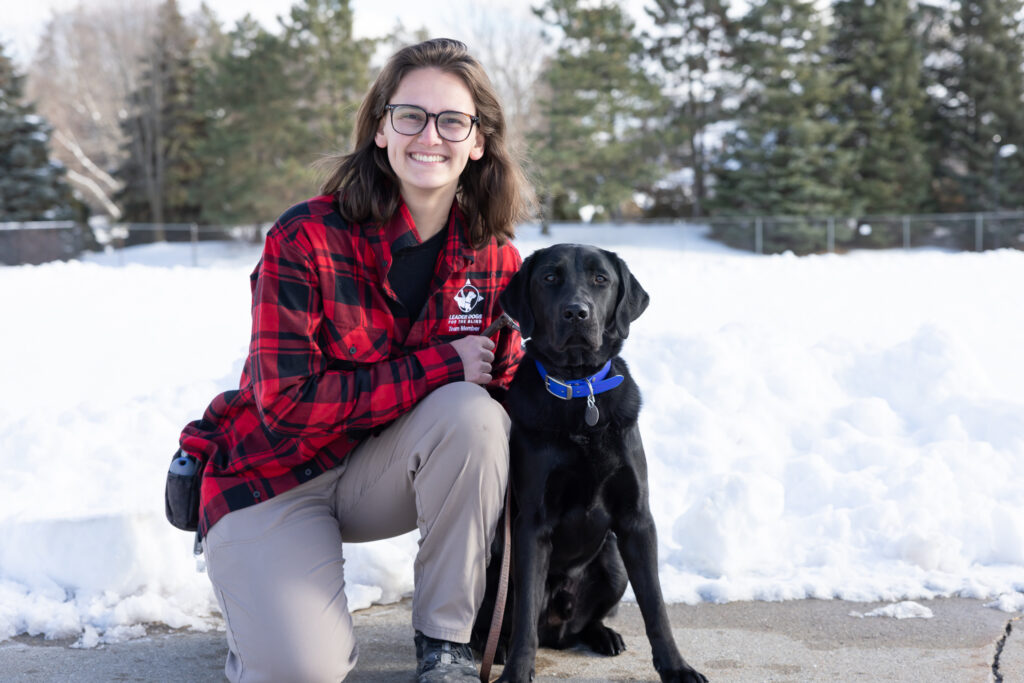 A woman with short brown hair smiles toward the camera. She kneels next to a black lab Leader Dog in harness. The dog looks toward the camera.