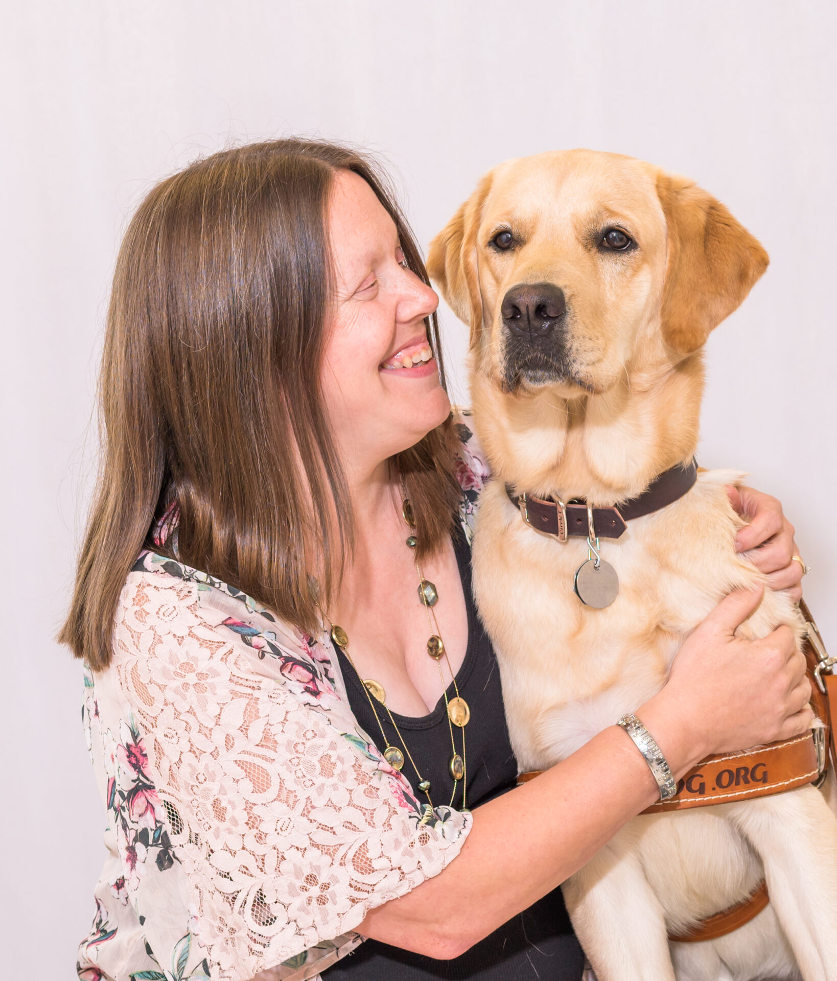 A woman with long brown hair smiles while holding a yellow Labrador guide dog in harness. The dog sits beside her, looking forward. The woman wears a floral lace kimono and layered necklace, and the background is a simple light backdrop.