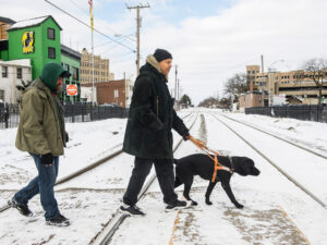 A person walks a dog along snowy train tracks while another individual trails behind in a winter coat.